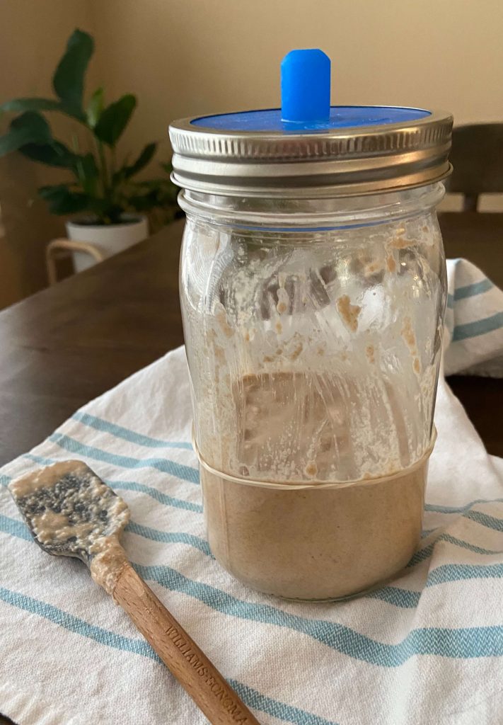 picture of sourdough starter in glass mason jar after being mixed sitting on kitchen towel and wooden spatula sitting next to the jar