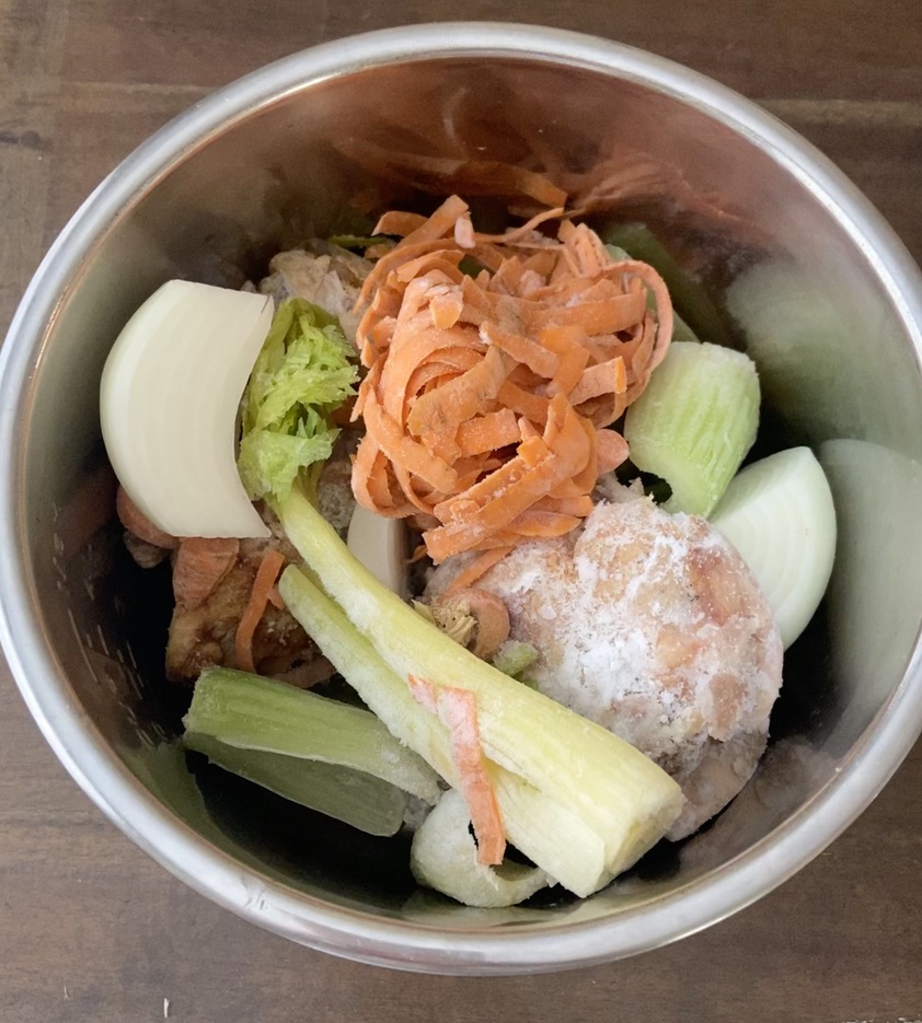 overhead image of frozen vegetables, celery, carrots and onions and frozen chicken bones in stainless steel container