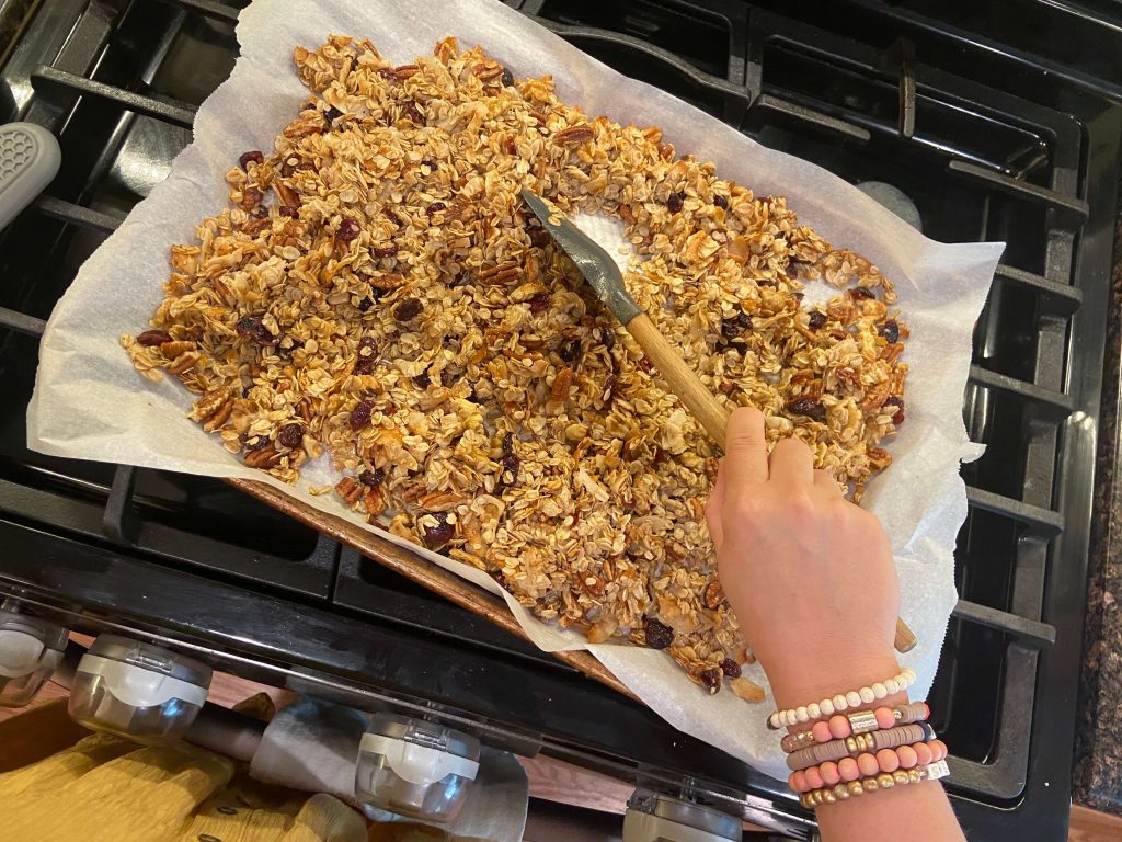 image of woman's hand stirring granola with wooden and silicon spatula on cookie sheet halfway through baking time