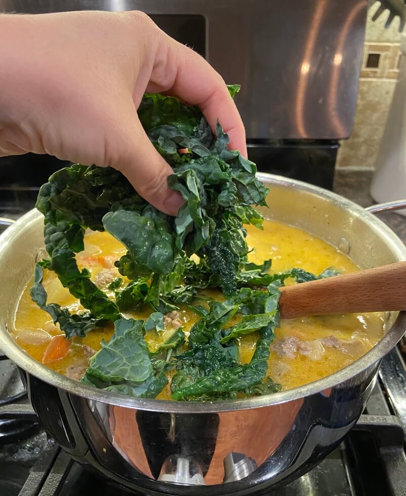 woman's hand dropping chopped kale into a pot of the best creamy tuscan sausage and kale soup zuppa toscana simmering on the stove