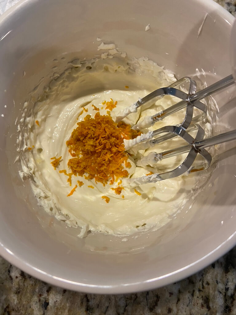 cream cheese in bowl with orange zest on top and hand mixer whipping ingredients together in white bowl