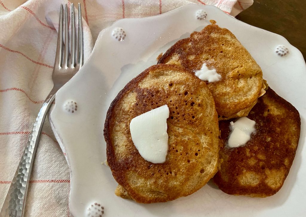 close up image of three cooked pancakes with butter on top on a white plate and fork next to it sitting on a table napkin