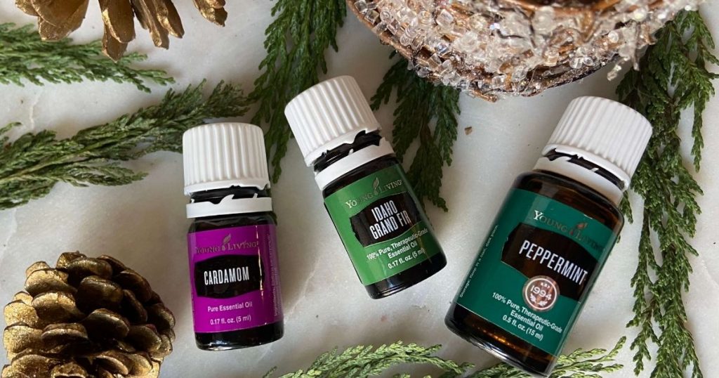 image of cardamom, idaho grand fir, and peppermint essential oils by young living on top of white marble table and christmas tree and pine cones and cedar in background