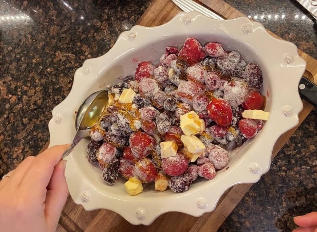 womans hand drizzling honey on frozen mixed berries mixed with flour and butter in white oval baking dish
