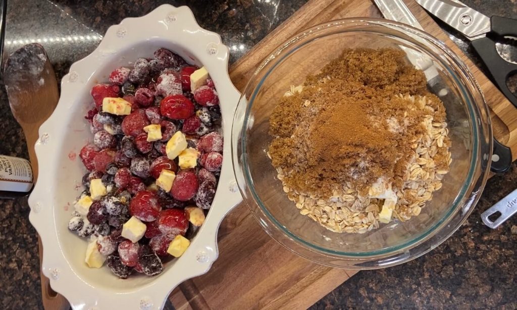 image of frozen mixed berries in white baking dish mixed with flour and butter and glass bowl with brown sugar oats cinnamon and flour sitting on cutting board