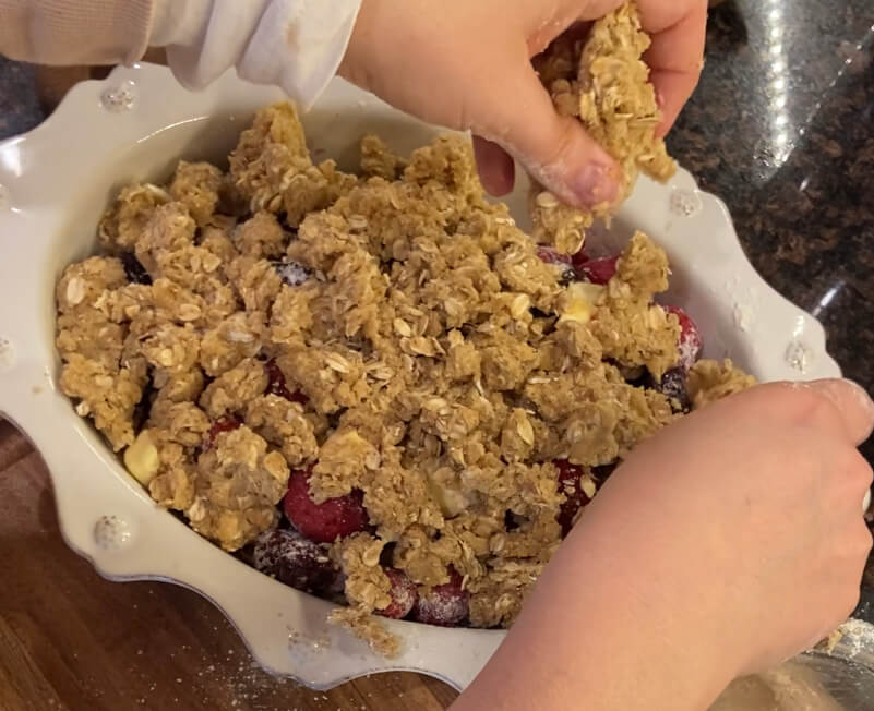 woman's hands sprinkling oat crumble topping on berry mixture in white baking dish