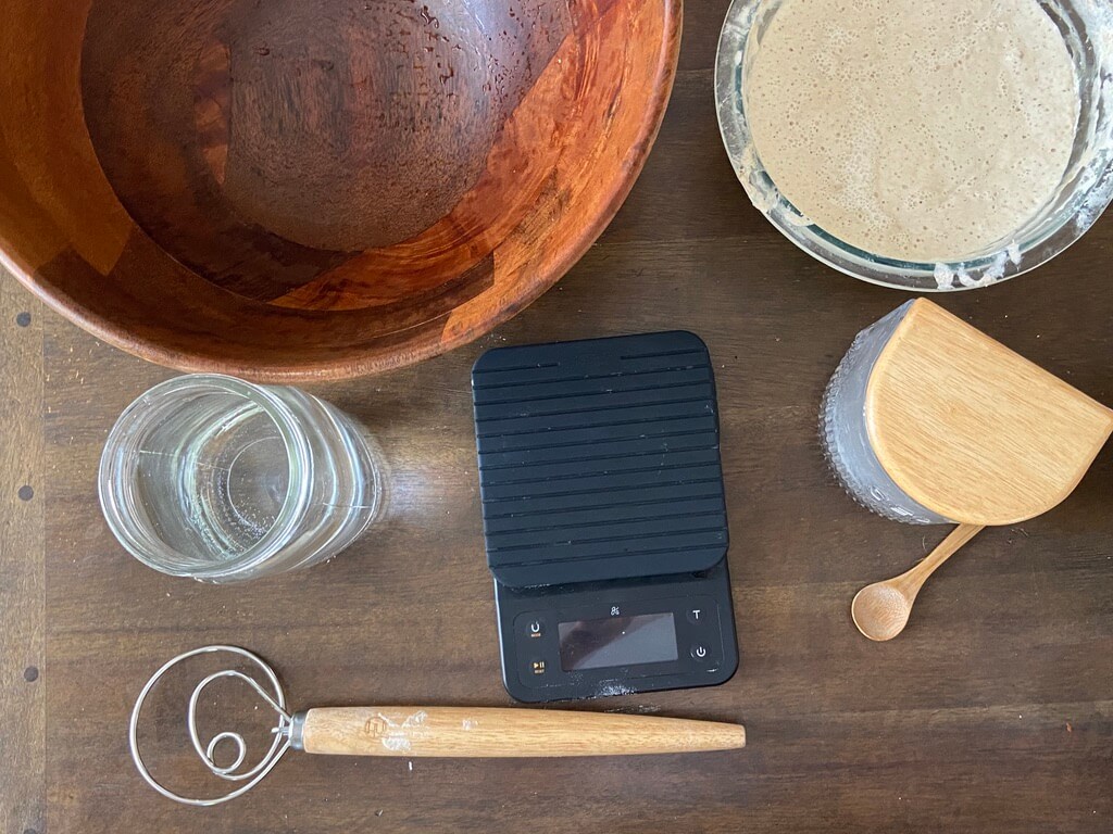 overhead image of wood bowl, glass bowl of sourdough starter, glass of water, dough whisk, kitchen scale, and salt jar with wooden spoon on wood table