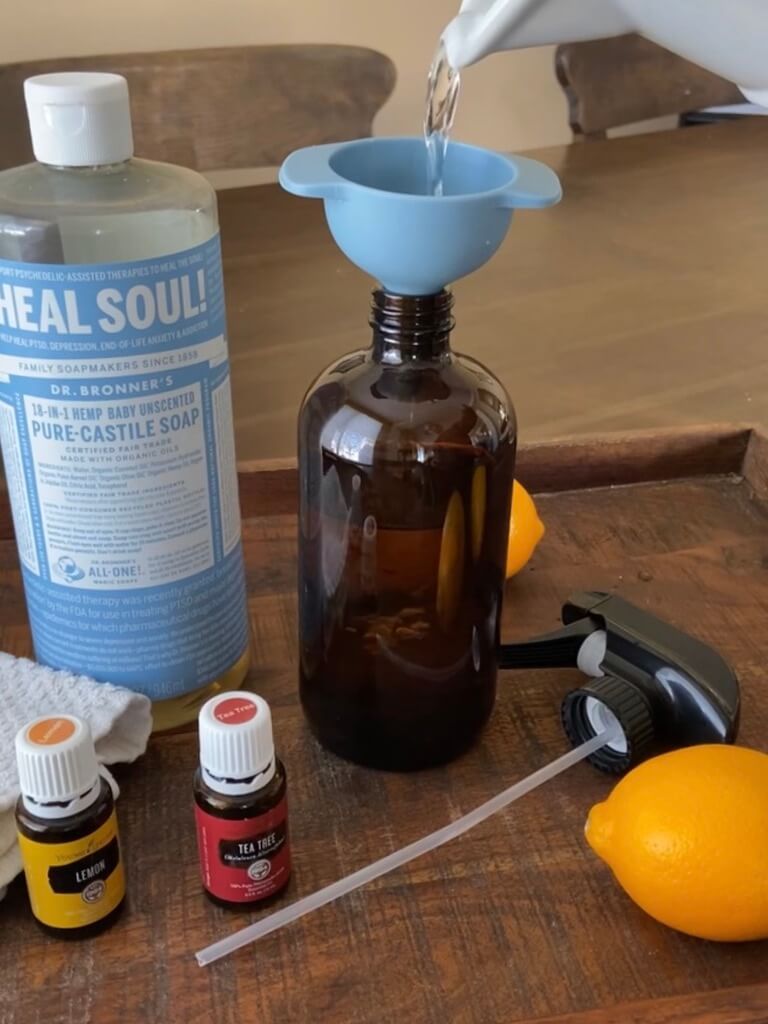 water pouring into an amber glass jar with a blue funnel and castile soap in background and essential oils in front all on wood tray