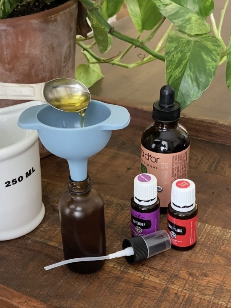 teaspoon of jojoba oil pouring into funnel and amber glass bottle with lavender and ylang ylang essential oils in background
